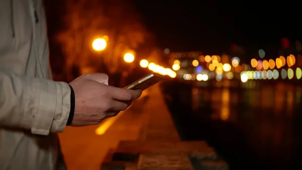 一个男人站在海滨, 晚上用手机在夜间灯光背景下, 背景模糊 — 图库照片