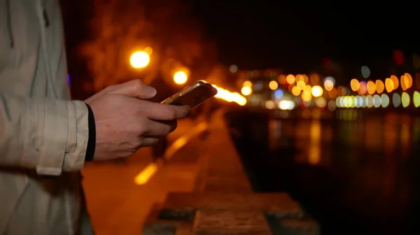 一个男人站在海滨, 晚上用手机在夜间灯光背景下, 背景模糊 — 图库照片