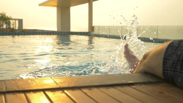 男はプールの端に座っているし、水に彼の足を濡らします。海の見える家の屋根の上の豪華なプール。夕日。水に太陽のまぶしさ. — ストック写真