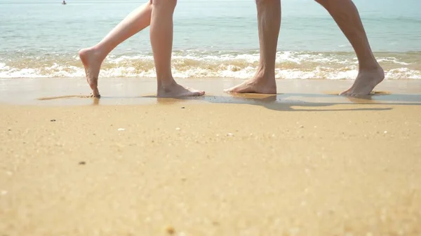 Una pareja está caminando por la playa en un día claro y soleado. se toman de la mano y se besan. los pies de hombres y mujeres caminando a lo largo de la orilla del mar — Foto de Stock