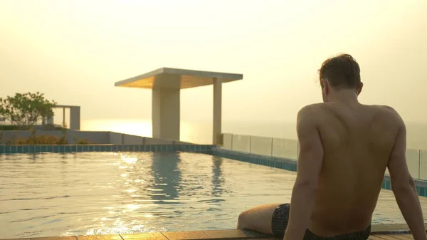 O homem senta-se na borda da piscina e molha os pés na água. uma piscina luxuosa no telhado da casa com vista para o mar. Ao pôr-do-sol. brilho solar na água . — Fotografia de Stock