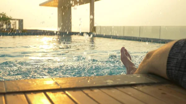 男はプールの端に座っているし、水に彼の足を濡らします。海の見える家の屋根の上の豪華なプール。夕日。水に太陽のまぶしさ. — ストック写真