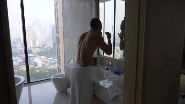 一个英俊的男人在浴室里应用剃刀, 从窗户到摩天大楼的景色. — 图库照片