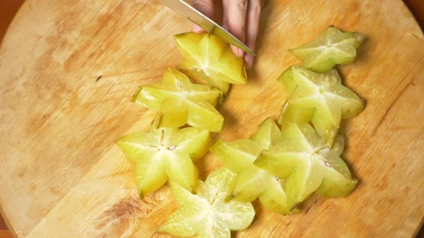 Frutas exóticas na mesa, as mãos femininas cortam uma carambola com uma faca em uma tábua de corte , — Fotografia de Stock