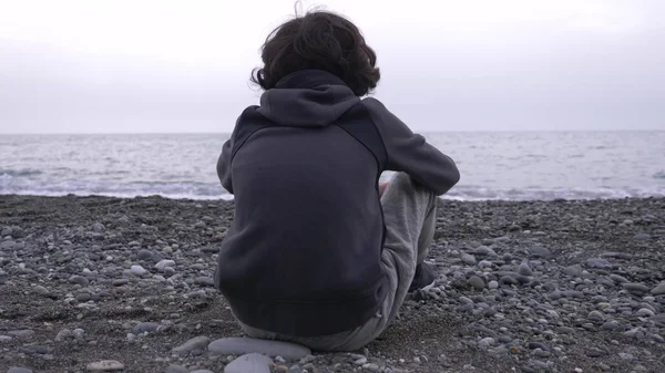 En ensam pojke sitter på stranden och kastar stenar på havet. — Stockfoto