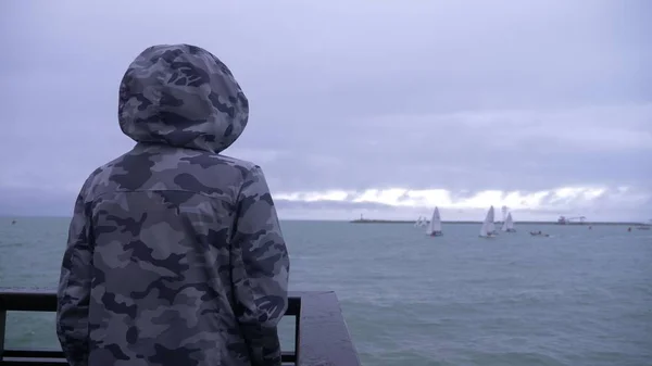 Jemand in Kapuzenjacke steht auf der Seebrücke und beobachtet die Segelboote, die aufs Meer fahren. — Stockfoto
