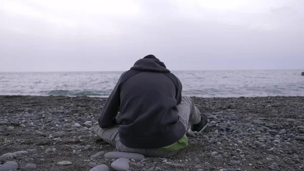 En ensam pojke sitter på stranden och kastar stenar på havet. — Stockfoto