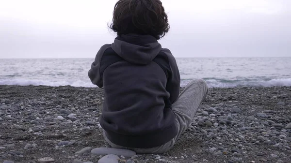 孤独な少年が海岸に座っているし、海に石を投げる. — ストック写真