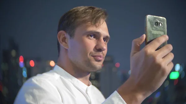 Joven y hermoso hombre usando un teléfono inteligente y bebiendo un cóctel, en una terraza del bar con vistas a los rascacielos por la noche. desenfoque de fondo . — Foto de Stock