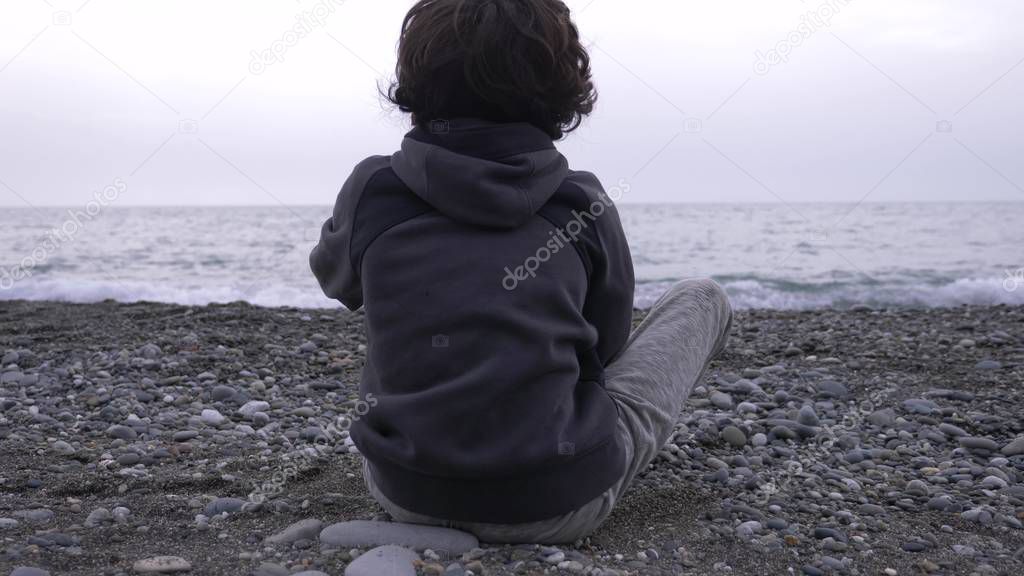 A lonely boy sits on the shore and throws stones at sea.