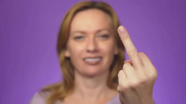 Young beautiful woman, blurred in the background, smiling. the focus of her hand showing middle fingers. colored background. — Stock Photo, Image