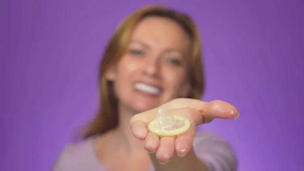 Ung vacker kvinna, suddig i bakgrunden, leende. i fokus är hennes hand som håller en öppen kondom, begreppet säker sex. Skydd mot Aids och födelsekontroll, Cologne bakgrunden — Stockfoto