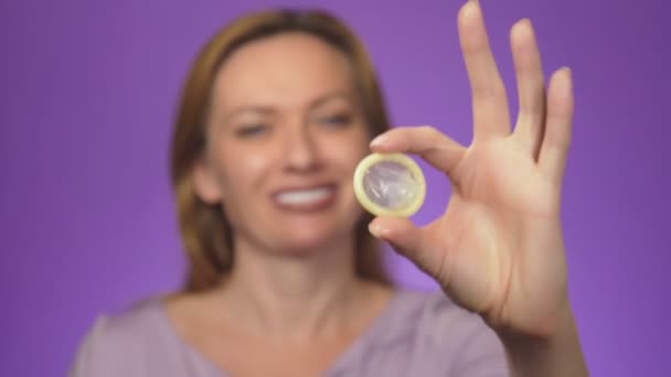 Joven mujer hermosa, borrosa en el fondo, sonriendo. en foco es su mano sosteniendo un condón abierto, el concepto de sexo seguro. Protección contra el SIDA y el control de la natalidad, fondo de color — Vídeos de Stock