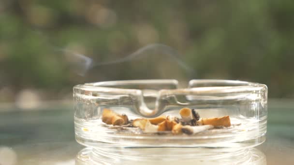 Rabos de cigarro de um cinzeiro de vidro em uma mesa de vidro ao ar livre . — Vídeo de Stock