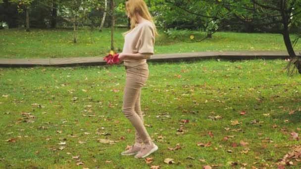 秋の公園を歩く若いブロンドの女性、彼女は落ちたもみじ紅葉を集めて — ストック動画