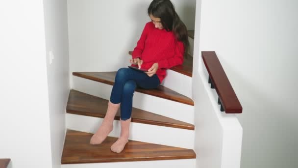 Красива дівчина в червоному светрі і махрові шкарпетки і джинси, сидячи на сходах, використовує планшет — стокове відео