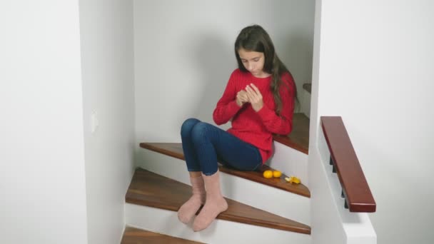 Beautiful girl in red sweater and jeans, sitting on the stairs, cleans tangerines and eats them. Christmas mood concept — Stock Video