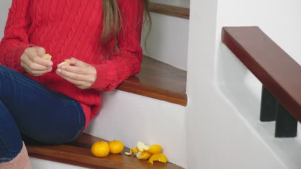 Красива дівчина в червоному светрі і джинсах, сидячи на сходах, очищає мандарини і їсть їх. Концепція різдвяного настрою — стокове відео