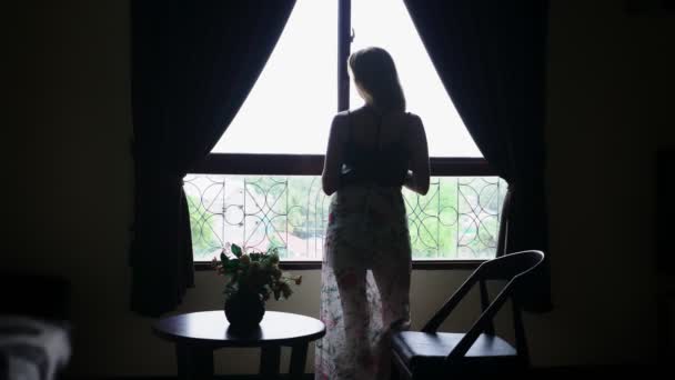 Silhouette. la fille va à une grande fenêtre avec un verre d'eau, boit de l'eau, regarde par la fenêtre et sourit . — Video