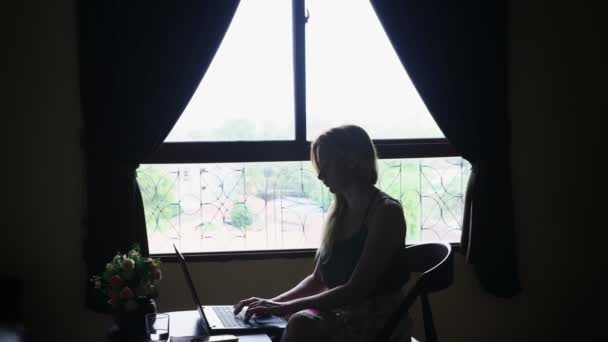 Silueta. chica se sienta en una silla junto a la ventana, utiliza su computadora portátil . — Vídeos de Stock