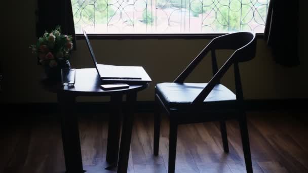 Siluetas. silla y mesa con portátil y flores en el fondo de una gran ventana. fuera de la vista del follaje verde de los árboles — Vídeo de stock