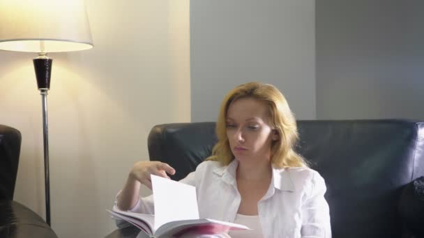 Uma mulher elegante em um terno branco se senta em uma grande cadeira de couro preto em sua sala de estar e lê um livro. espaço de cópia — Vídeo de Stock