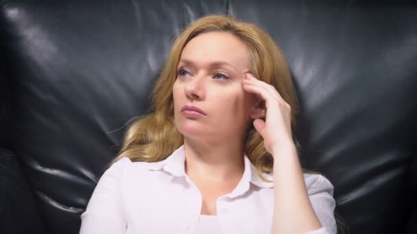 一个穿着白色西装的优雅女人 坐在一张黑色的大皮椅上 想着什么 看着镜头 微笑着 复制空间 — 图库视频影像