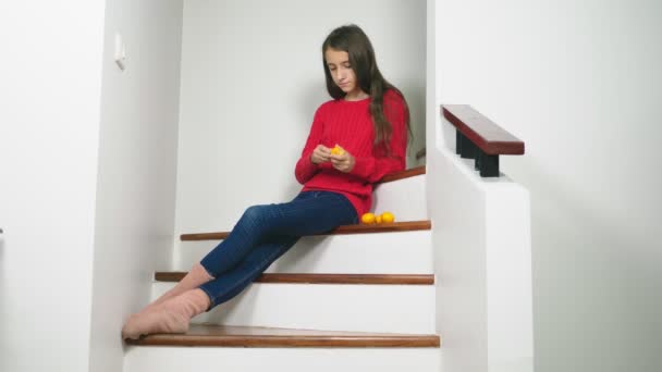 Hermosa chica en suéter rojo y jeans, sentada en las escaleras, limpia mandarinas y se las come. Concepto de humor de Navidad — Vídeos de Stock
