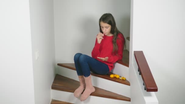 Hermosa chica en suéter rojo y jeans, sentada en las escaleras, limpia mandarinas y se las come. Concepto de humor de Navidad — Vídeo de stock