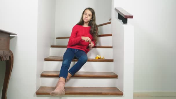 Hermosa chica en suéter rojo y jeans, sentada en las escaleras, limpia mandarinas y se las come. Concepto de humor de Navidad — Vídeos de Stock