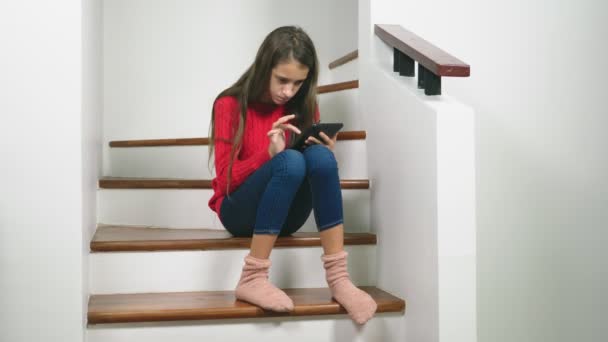 Hermosa chica en un suéter rojo y terry calcetines y vaqueros, sentado en las escaleras, utiliza la tableta — Vídeo de stock