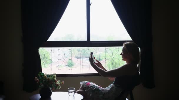 Silhouette. ragazza si siede su una sedia contro la finestra, usa il telefono e beve acqua da un bicchiere — Video Stock