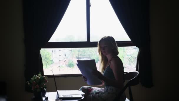 Silueta. chica se sienta en una silla junto a la ventana, utiliza su computadora portátil, mirando a través de documentos de papel — Vídeos de Stock