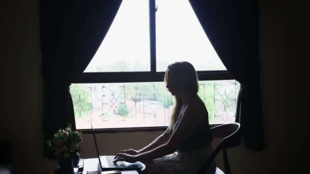 Silueta. chica se sienta en una silla junto a la ventana, utiliza su computadora portátil . — Vídeos de Stock