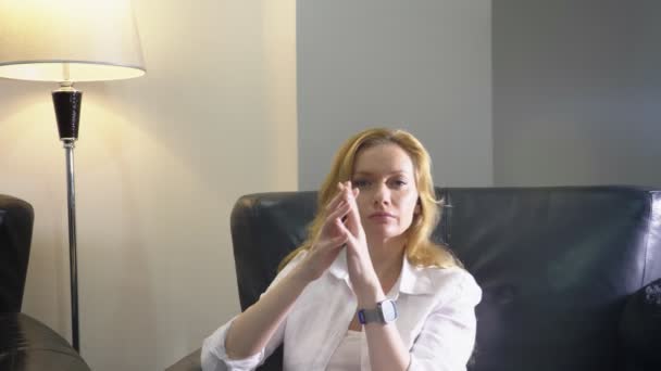 Крупным планом. Элегантная женщина в белом костюме сидит в большом черном кожаном кресле, смотрит в камеру и показывает руки жестом превосходства, складывая пальцы шпилем. копировальное пространство — стоковое видео