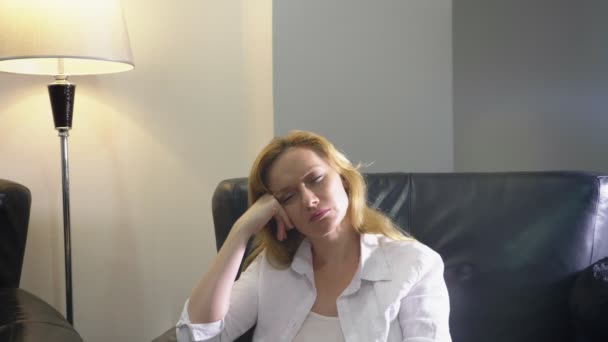 一个穿着白色家居服的优雅女人坐在客厅里的一把黑色的大皮椅上 正在想着什么 看着镜头 面带微笑 复制空间 — 图库视频影像