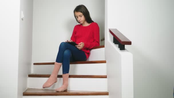Hermosa chica en un suéter rojo y terry calcetines y vaqueros, sentado en las escaleras, utiliza la tableta — Vídeo de stock