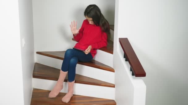 Vacker flicka i en röd tröja och terry strumpor och jeans, sitter på trappan, använder tabletten — Stockvideo