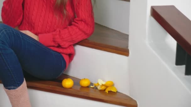 Fata frumoasa in pulover rosu si blugi, asezata pe scari, curata mandarine si le mananca. Conceptul de Crăciun — Videoclip de stoc