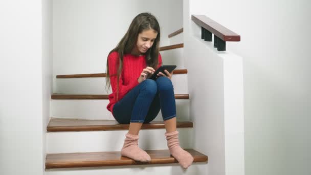 Hermosa chica en un suéter rojo y terry calcetines y vaqueros, sentado en las escaleras, utiliza la tableta — Vídeos de Stock