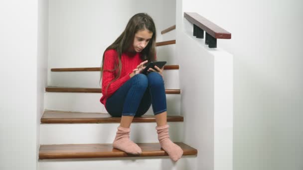 Bella ragazza in un maglione rosso e calzini di spugna e jeans, seduto sulle scale, utilizza il tablet — Video Stock