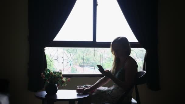 Silueta. chica se sienta en una silla contra la ventana, usa su teléfono y bebe agua de un vaso — Vídeos de Stock