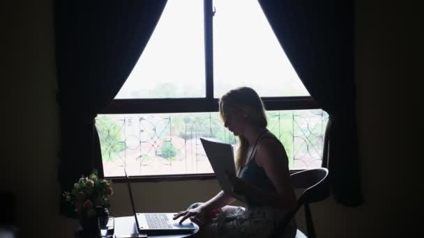 Silueta. chica se sienta en una silla junto a la ventana, utiliza su computadora portátil, mirando a través de documentos de papel — Vídeo de stock