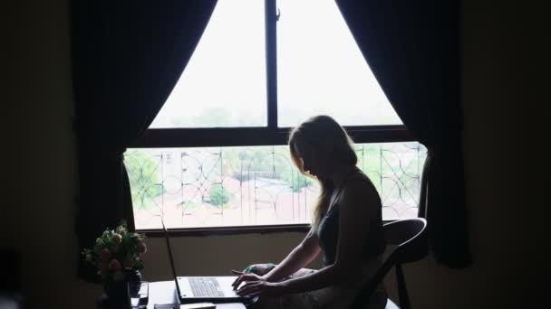 Silueta. chica se sienta en una silla junto a la ventana, utiliza su computadora portátil . — Vídeos de Stock