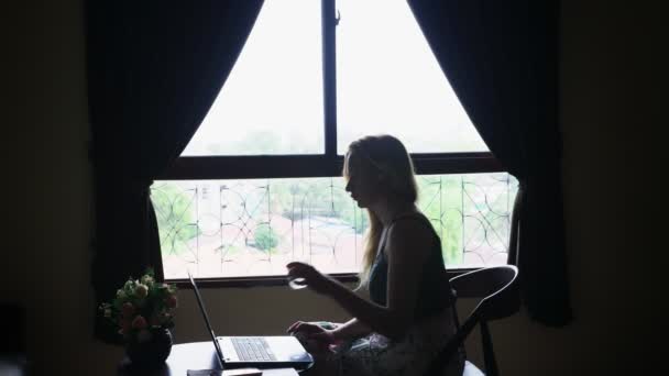 Silhouette. fille s'assoit sur une chaise près de la fenêtre, utilise son ordinateur portable . — Video