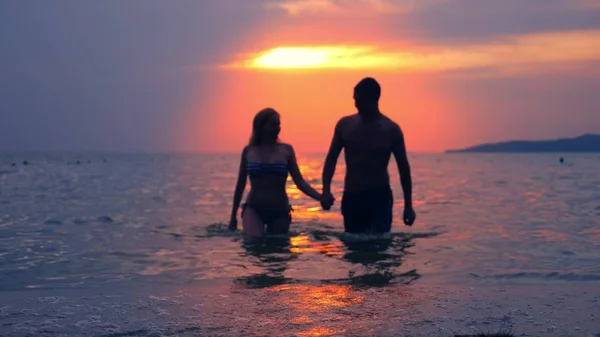Siluetas, pareja, hombre y mujer fuera del mar tomados de la mano contra el fondo del paisaje marino, rojo dramático atardecer en el mar. el sol tiñe el mar de rojo . — Foto de Stock