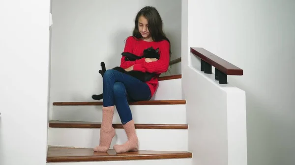 美丽的少女在红色毛衣和特里袜子和牛仔裤, 坐在楼梯上, 微笑着, 抱着一只小猫在她的腿上 — 图库照片