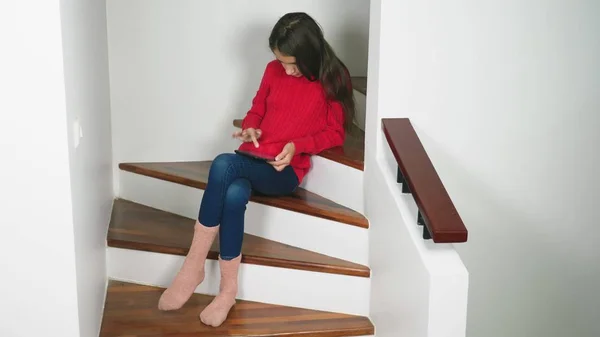 Mooi meisje in een rode trui en sokken van terry en jeans, zittend op de trap, maakt gebruik van de tablet — Stockfoto