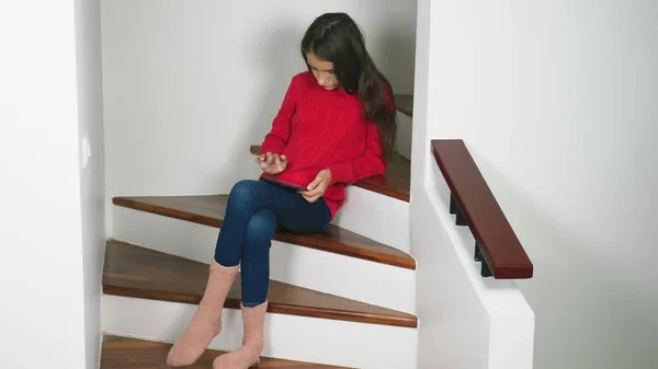 Schönes Mädchen in rotem Pullover und Frottee-Socken und Jeans, sitzt auf der Treppe und benutzt das Tablet — Stockfoto