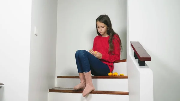 Schönes Mädchen in rotem Pullover und Jeans, sitzt auf der Treppe, putzt Mandarinen und isst sie. Konzept zur Weihnachtsstimmung — Stockfoto